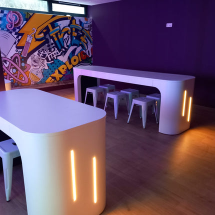 Modern white curved battery powered tables with off centre integrated warm white 3000K LED light lines. 4mm COB LED strips used to add ambience to creative graffiti wall leisure room. Installed within David Lloyds, Ucle in Belgium, a high profile fitness centre.