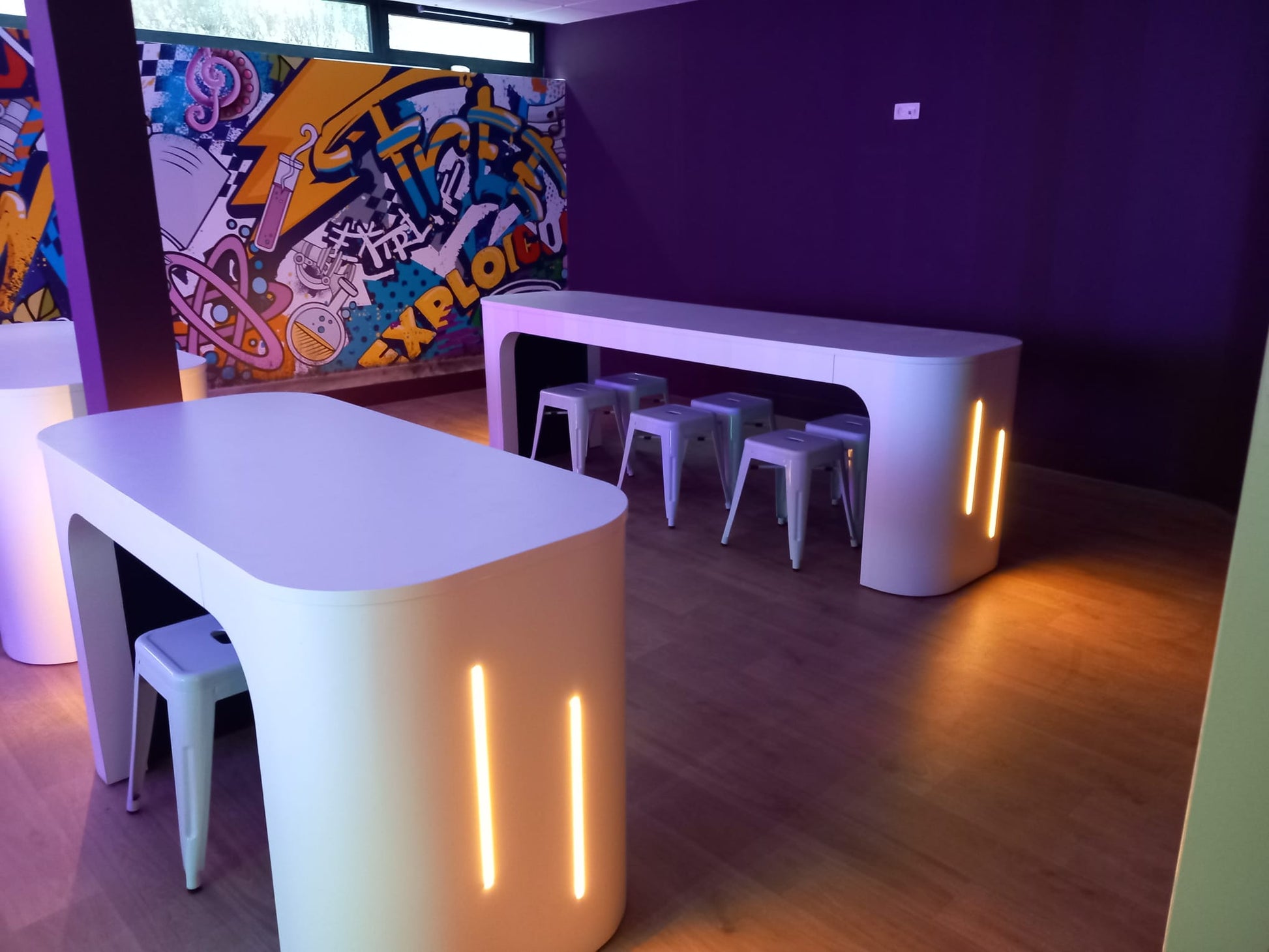 Modern white curved battery powered tables with off centre integrated warm white 3000K LED light lines. 4mm COB LED strips used to add ambience to creative graffiti wall leisure room. Installed within David Lloyds, Ucle in Belgium, a high profile fitness centre.
